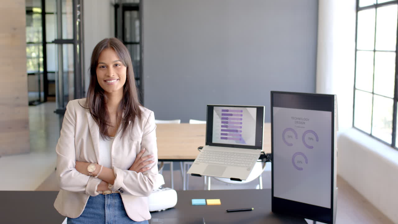 Standing with arms crossed, smiling businesswoman in office with laptop and charts