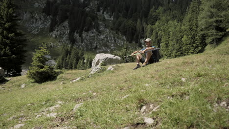 Die-Kamera-Fährt-Nach-Vorne-Auf-Den-Wanderer-Zu,-Der-Mit-Wanderstöcken-In-Den-Händen-Auf-Grünem-Gras-Am-Boden-Sitzt