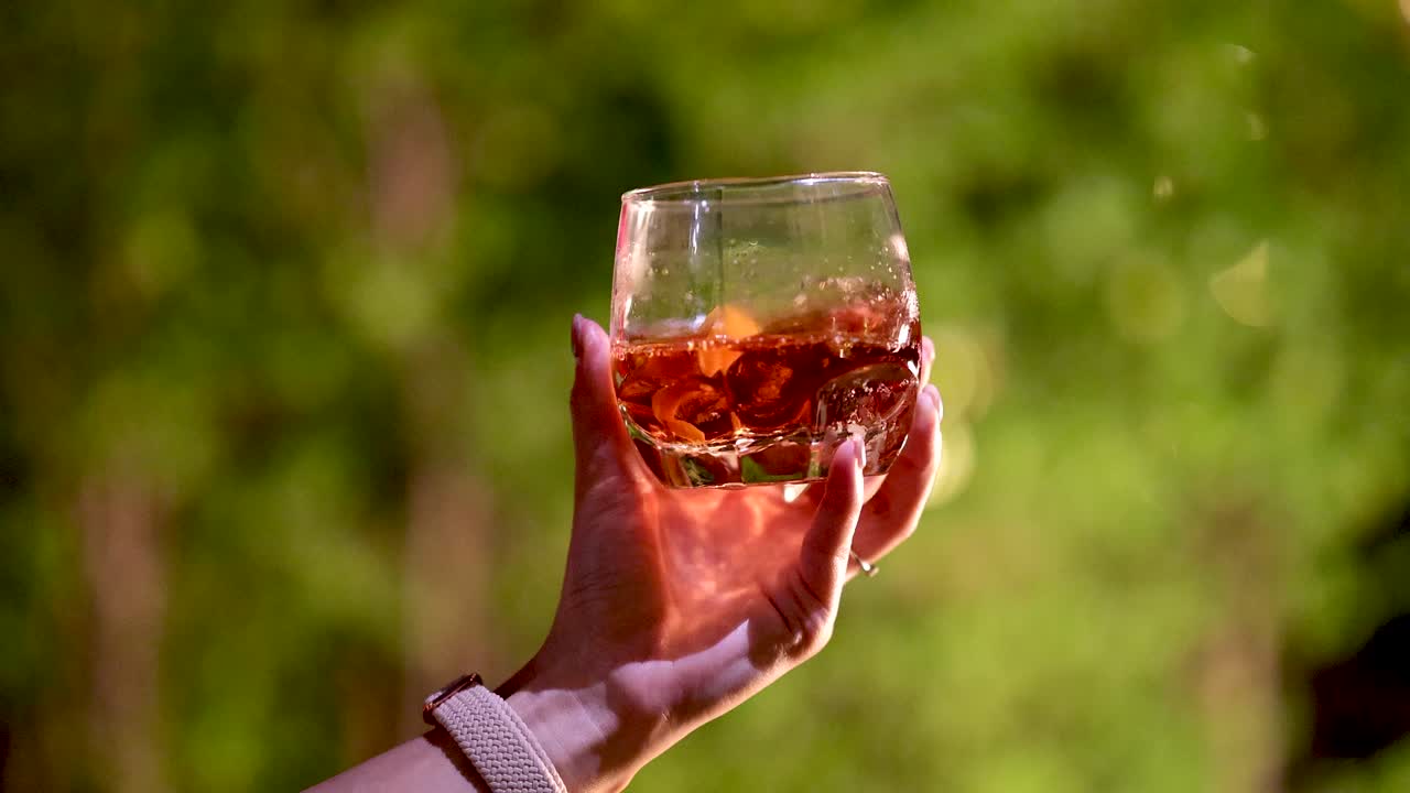 Person lifts Negroni cocktail glass against sunlit greenery, warm natural lighting, shallow depth