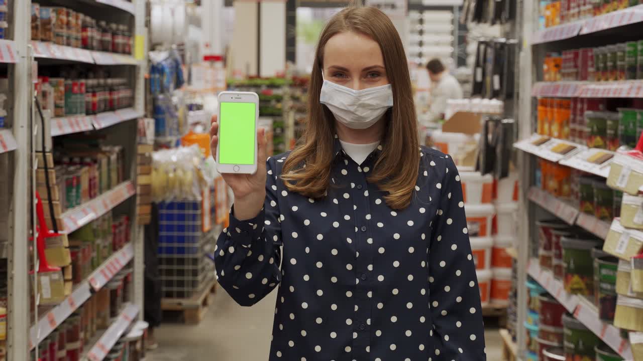 Woman wearing a protective mask, pointing at the green screen of a smartphone, looks at the camera in a hardware store