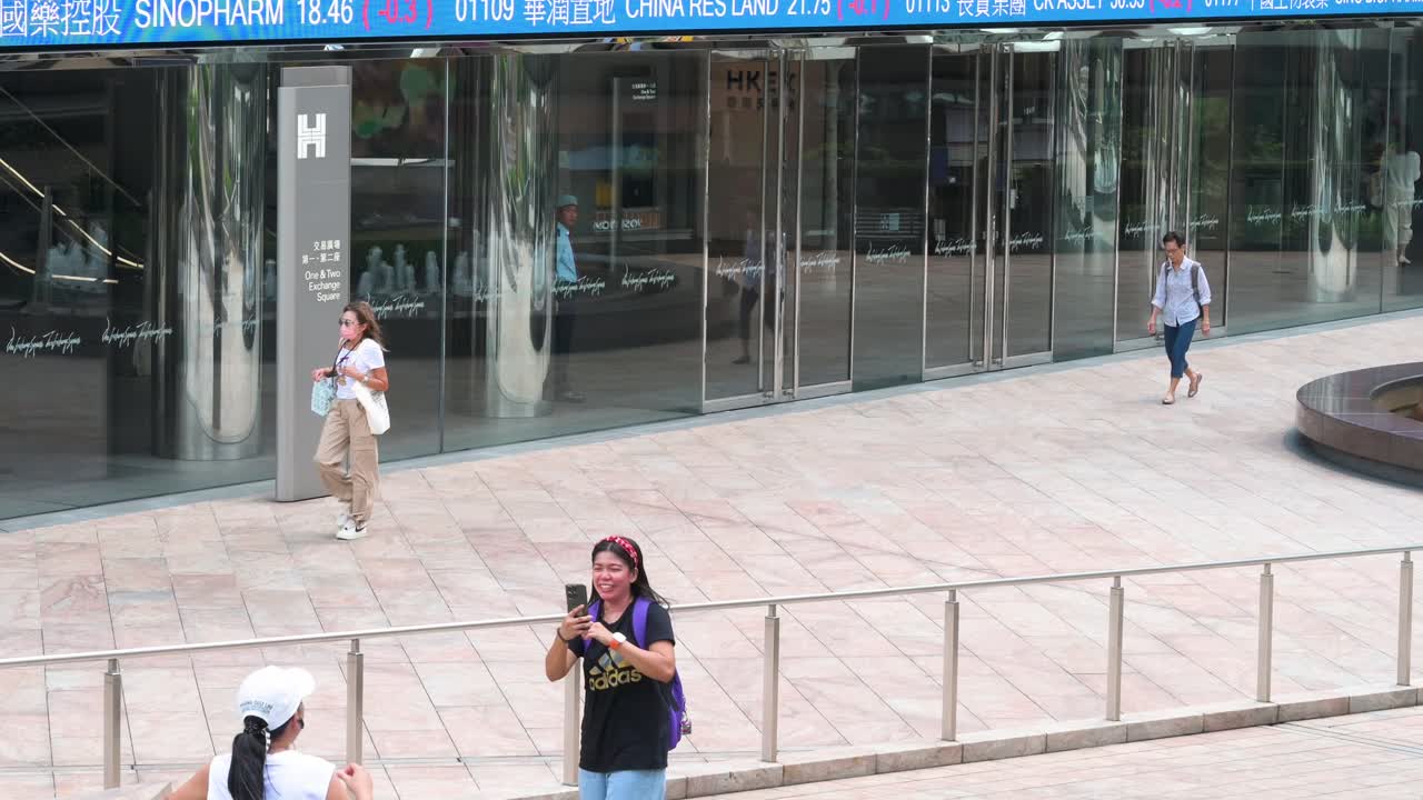 People are busy taking pictures as a moving screen in the background highlights negative stock ticker symbols at Exchange Square, the site of the Hong Kong Stock Exchange in the financial district.