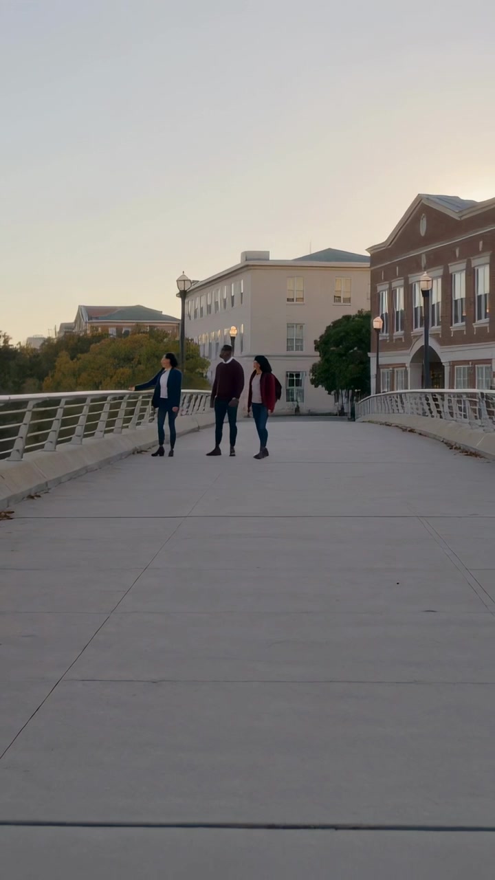 Video verticale: Trio che cammina su un ponte del campus al tramonto, il più a sinistra che guarda oltre la ringhiera, giacca rossa
