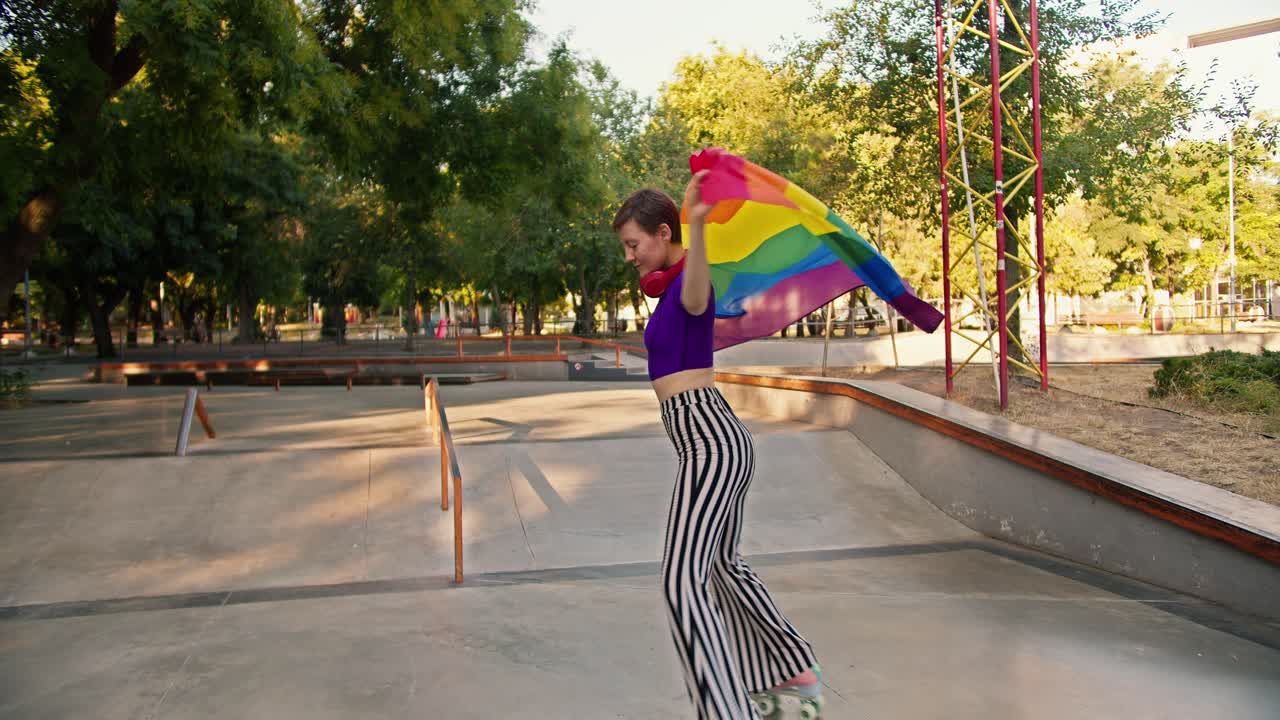 una chica feliz con un corte de pelo corto en una camiseta púrpura y pantalones a rayas monta patines rosados en un parque de patinaje y se envuelve en la bandera lgbt