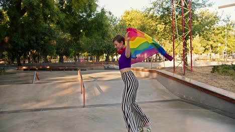 una chica feliz con un corte de pelo corto en una camiseta púrpura y pantalones a rayas monta patines rosados en un parque de patinaje y se envuelve en la bandera lgbt