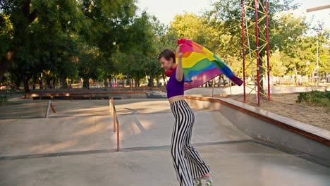 a happy girl with a short haircut in a purple tank top and striped pants rides pink roller skates in a skate park and wraps herself in the lgbt flag