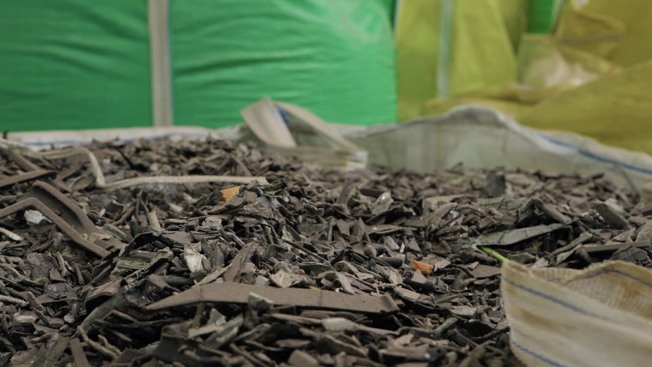 Large bulk bag overflowing with shredded dark mixed plastic strips and fragments inside recycling facility