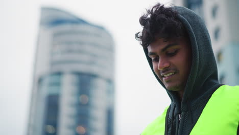 young male worker in neon reflective vest and dark hoodie standing outdoors in urban area, looking away with serious focused expression, modern glass buildings blurred in background on cloudy day