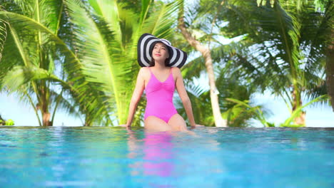 young female tourist sitting on the edge of a swimming pool wearing a pink one-piece swimwear and a floppy hat with palm trees blurred in the background