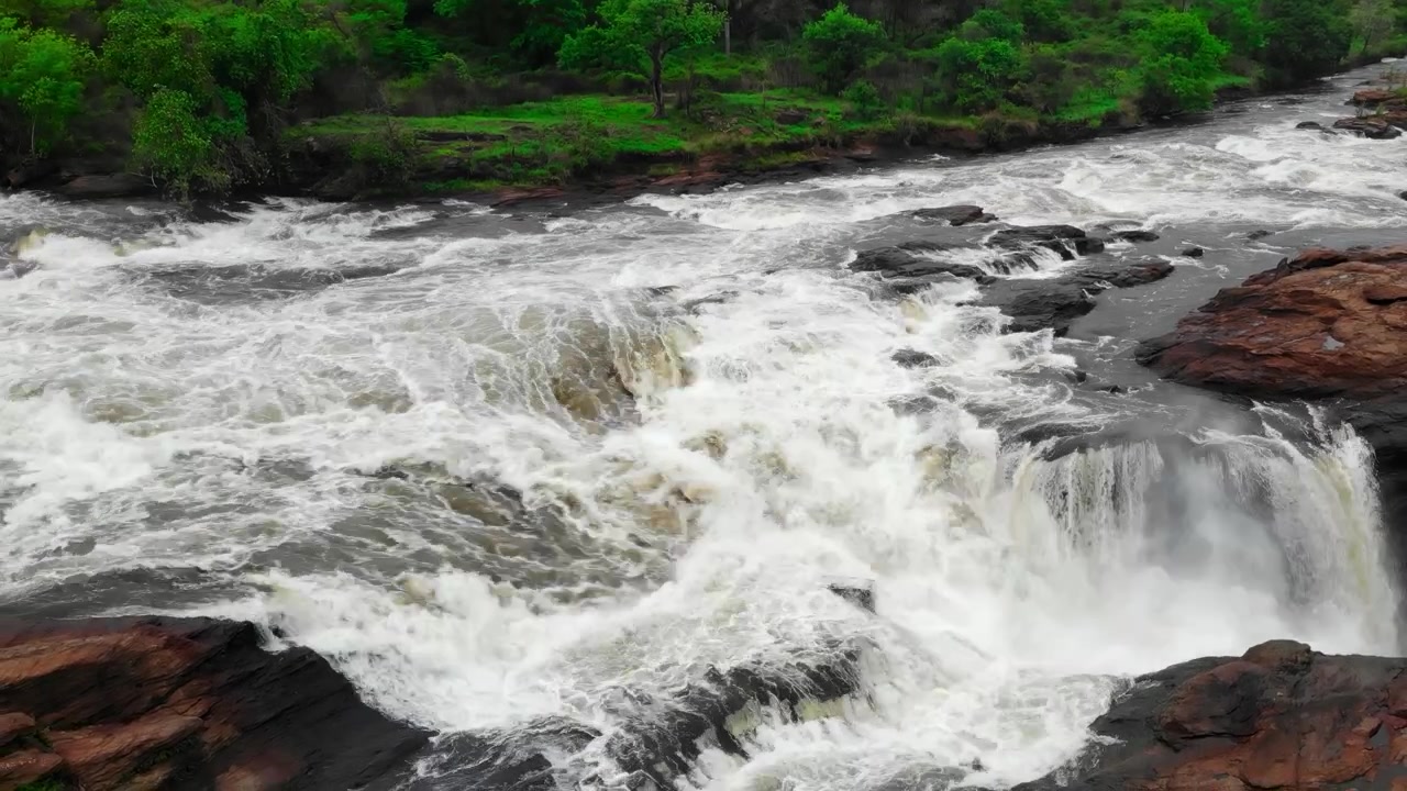 A wide view of a fast-moving waterfall and rapids flowing through dark rocks beside a lush green forest in natural daylight, showing turbulent water, foam, and scenic wilderness.