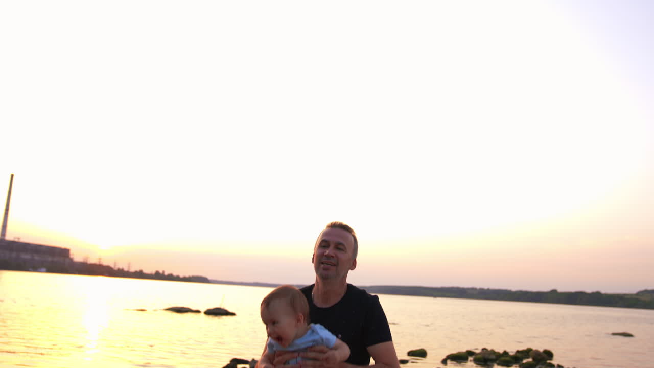 Happy father and son playing cheerfully at the waterfront. Dad is tossing his baby boy and he is laughing excitedly. River at backdrop.