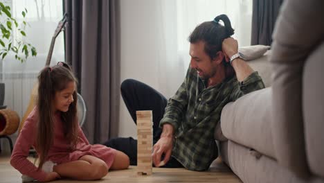 Una-Niña-Morena-Con-Un-Vestido-Rosa-Juega-Con-Su-Padre,-Un-Hombre-Moreno-Con-Una-Camisa-A-Cuadros-Verde,-En-El-Juego-De-Mesa-Jenga-Mientras-Está-Sentado-En-El-Suelo-Apoyado-En-Un-Sofá-Gris-En-Un-Apartamento-Moderno.