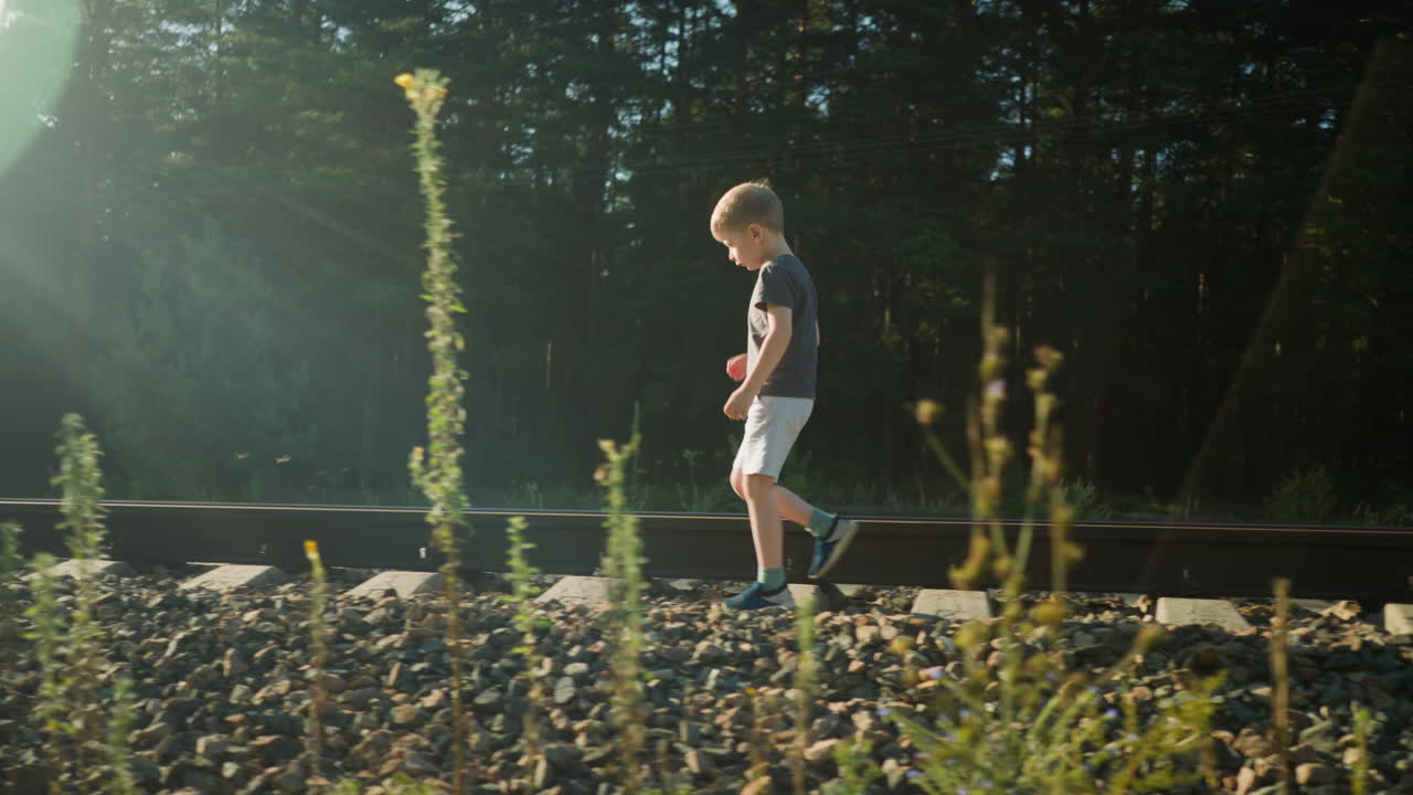 Young boy in dark shirt and light shorts walking carefully along gravel beside rail track as sunlight filters through forest, creating lens glow and long shadows over rural landscape