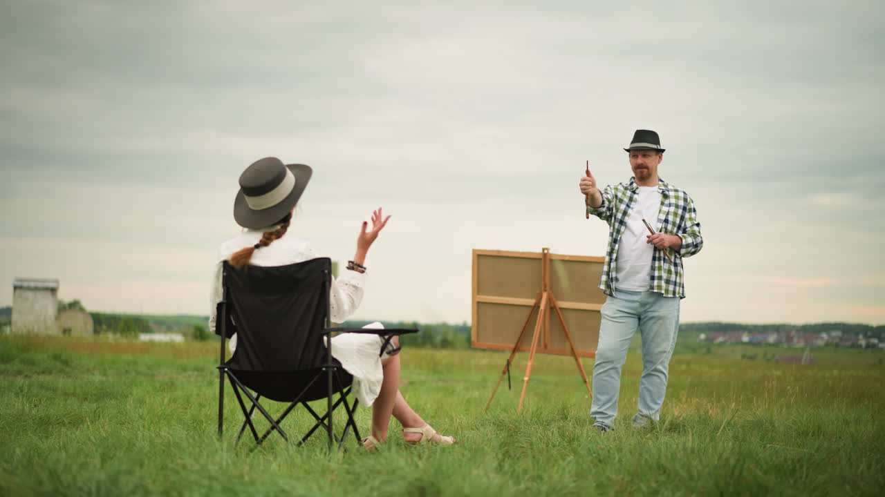 체크 된 셔츠와 청바지를 입은 예술가는 잔디 에 의자에 앉아있는 여성을 향해 브러쉬를 가리키며 미소 짓고 있습니다. 큰 보드는 배경에 설정되어 있습니다.