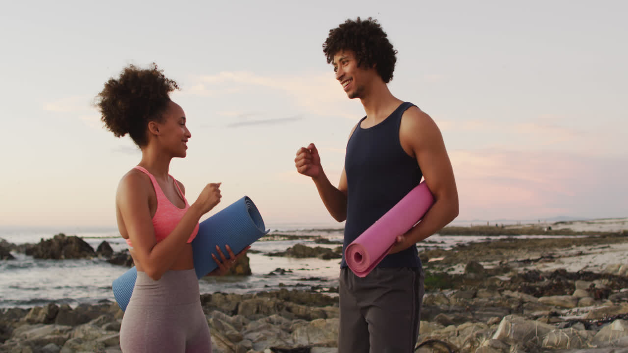 una feliz pareja biracial hablando con esteras de yoga en la playa