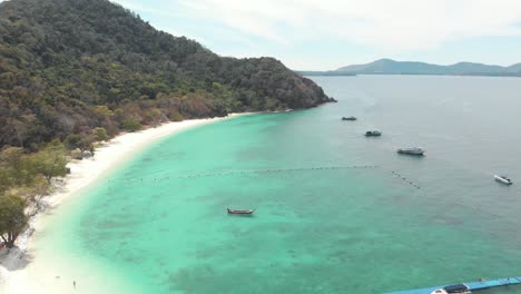 lush green steep hill and sandy shoreline encircling shallow turquoise sea of koh hey with moored boats in thailand - aerial fly-over rotating orbit shot