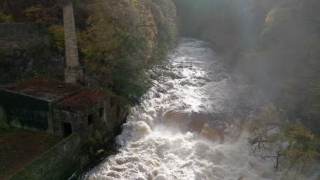 Drohnenaufnahmen-Fliegen-Direkt-Auf-Einen-Schnell-Fließenden-Fluss-Und-Wasserfall-Zu,-Umgeben-Von-Alten-Gebäuden-Und-Einem-Wald-Aus-Herbstlichen-Laub--Und-Nadelbäumen