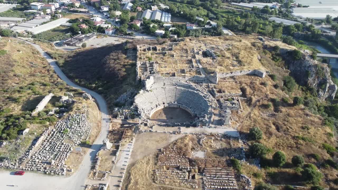 Aerial drone view showing semi circular amphitheater with surrounding stone ruins of ancient city Myra in Antalya Province under bright daylight across dry terrain mixed with patches of greenery