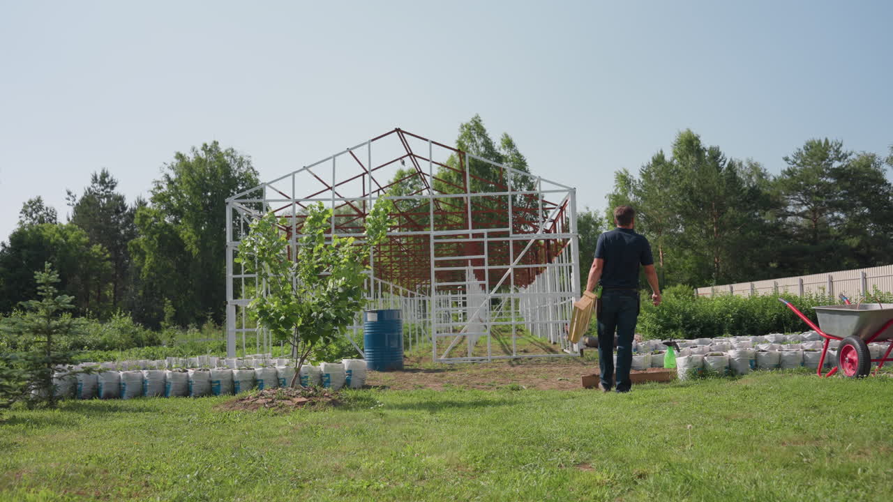 Rear view horticulturist walking toward farm carrying crate wiping forehead while looking over crops near metal frame structure and wheelbarrow on sunny rural yard