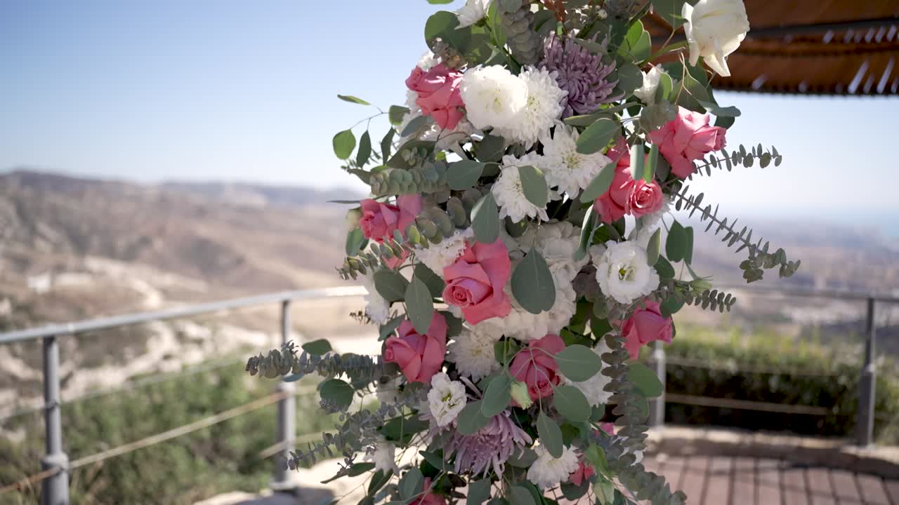 A flower arrangement hanging from a balcony with a view of hills and valleys. The flowers include pink roses and white blooms. It is a sunny day