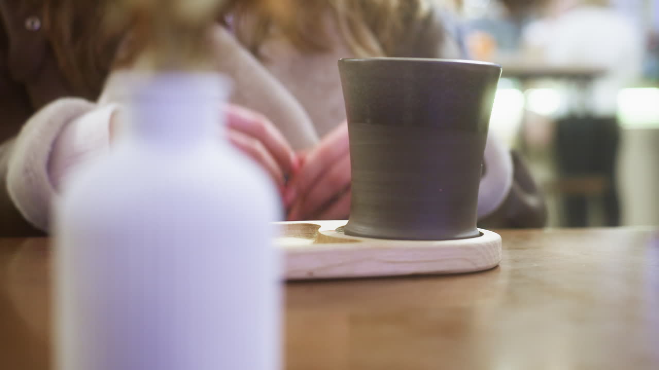 Close-up of hand in brown shearling jacket gently lifting cup of coffee at cozy cafe table. Soft lighting creates warm atmosphere, capturing peaceful moment of relaxation and comfort in cafe