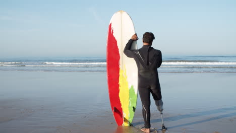 vista posteriore di un surfista maschio in muta appoggiata alla tavola da surf e in piedi di fronte al mare guardando le onde 1