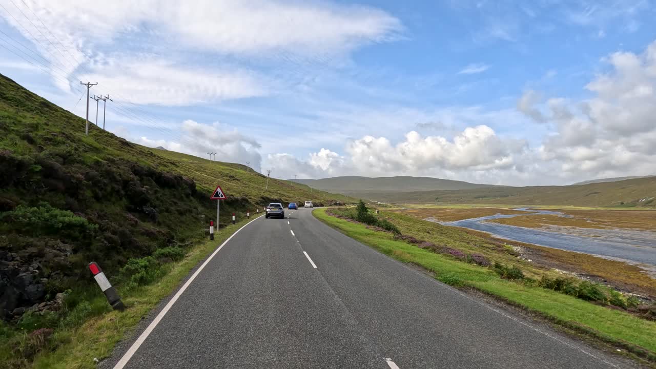 A vehicle travels a winding rural road beside hills and water under bright daylight, captured from a moving perspective with clear skies and natural landscape