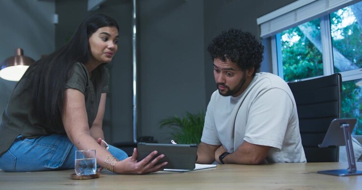 Couple sitting at home office table woman bringing tablet, gesturing to discuss