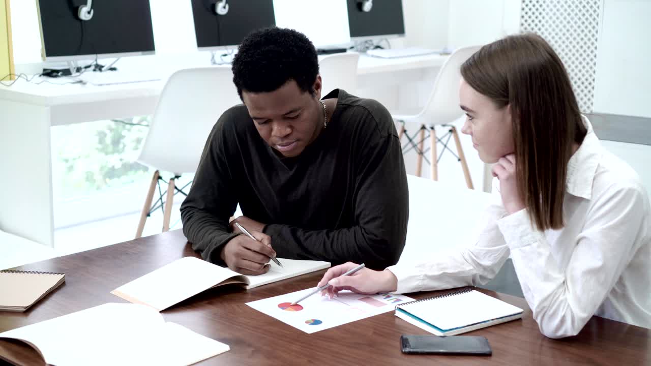 dos estudiantes de marketing trabajando juntos en un proyecto. joven africano y mujer caucásica sentados en un escritorio y discutiendo un documento con datos estadísticos