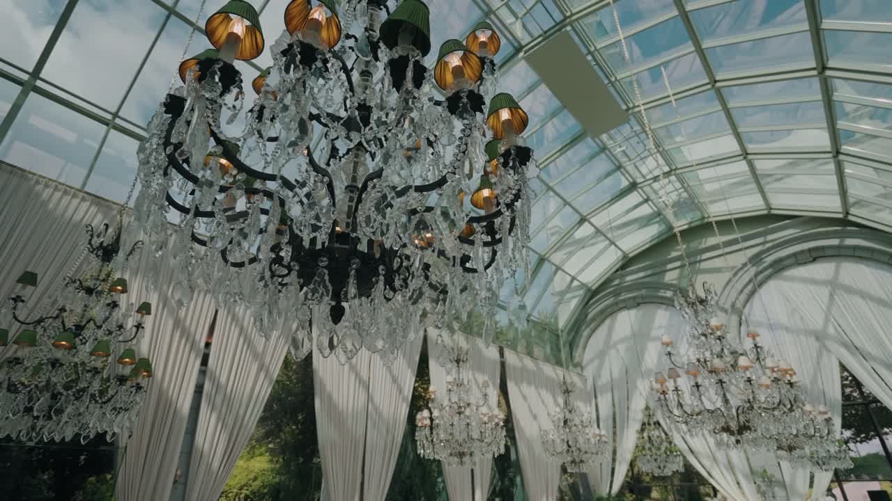 Close up of ornate crystal chandeliers inside a bright glass-roofed ballroom or event venue