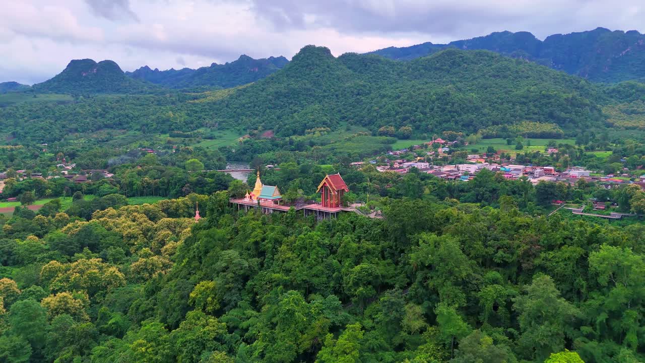 Aerial orbit around a golden temple perched on a mountain summit near Sangkhlaburi, Thailand, with views of the forest, village, river, and dramatic limestone mountains