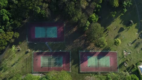 Drone-with-camera-down-slowly-descending-towards-3-tennis-courts-with-nature-around-in-public-park,-sports,-health,-leisure,-in-4K-resolution