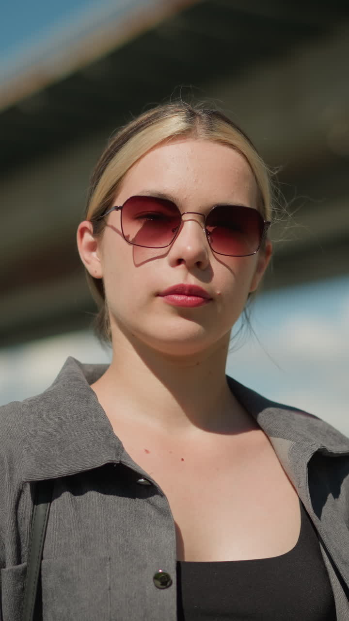 женщина в серой рубашке и солнцезащитных очках наклоняет голову слегка вправо, улыбаясь, она выглядит сосредоточенной, находясь на открытом воздухе, с размытым фоном с мостом и ярким небом