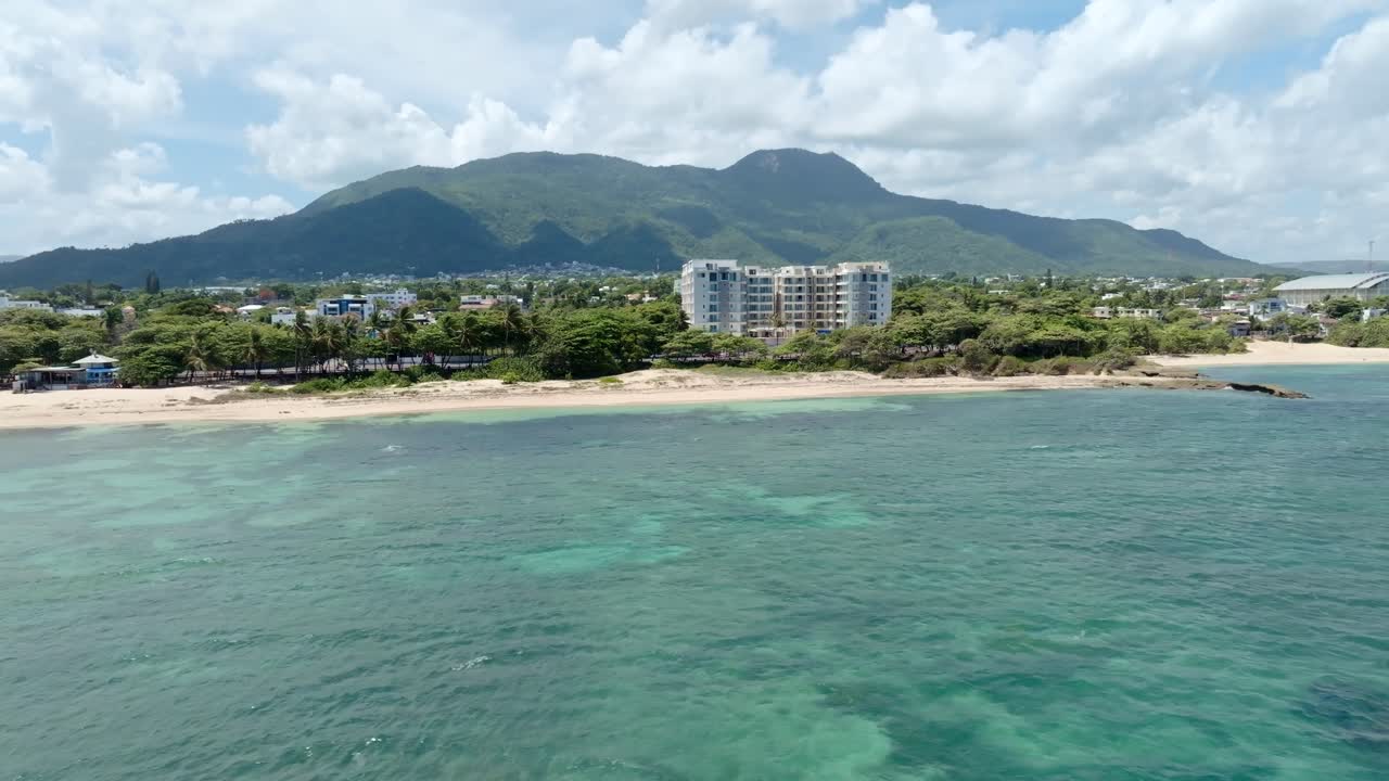 Aerial wide shot showing Sandy beach of puerto plata in Dominican Republic. Waterfront houses and apartment compel hotel. Green mountains in distance