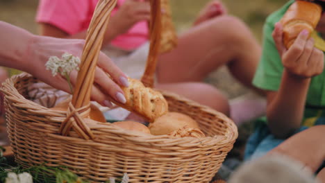 close-up of family seated outdoors picking fresh pastries from a picnic basket, young boy in green shirt quickly eats his choice while others select their treats, cozy picnic moment