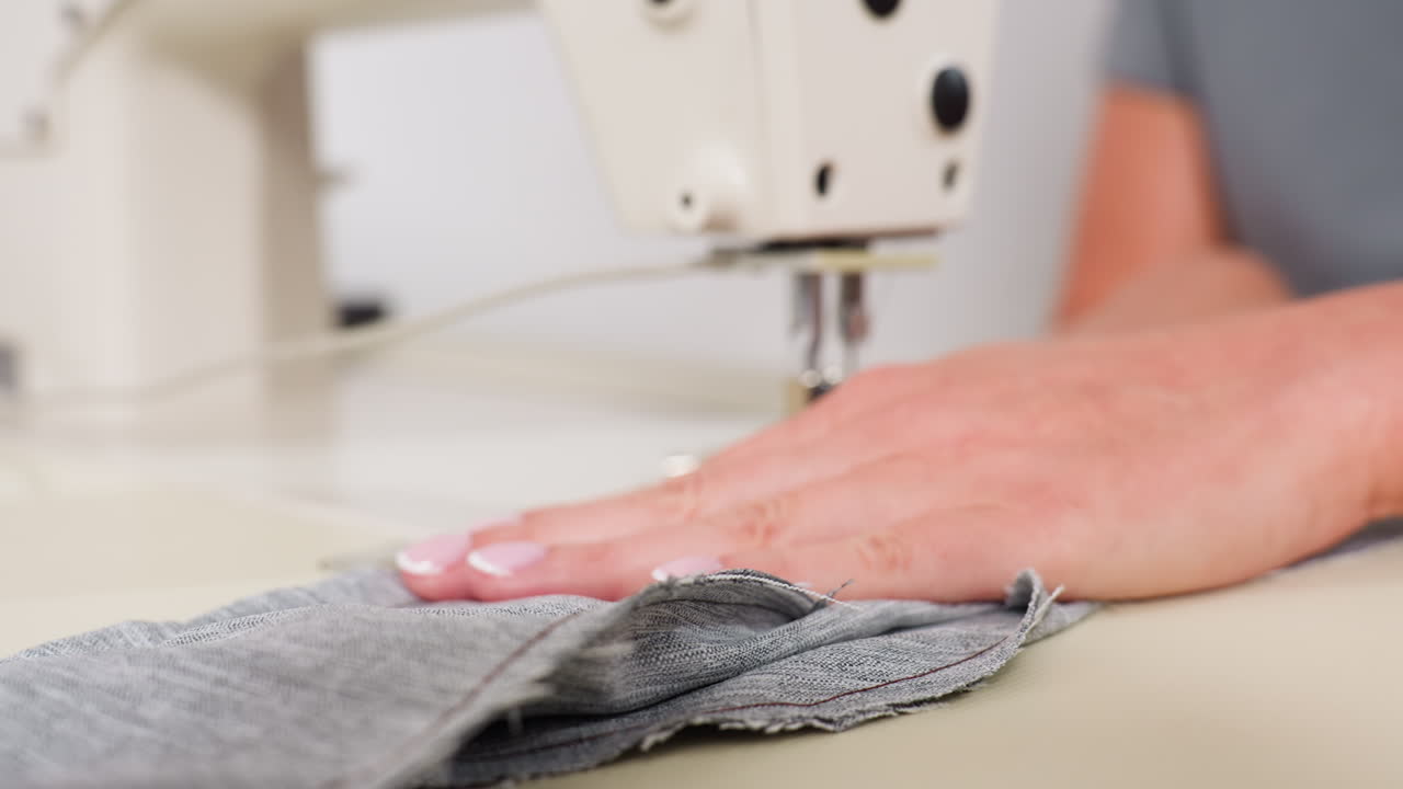 Hand view of design originator carefully sewing apparel through needle, adjusting fabric with precise handling, focusing on stitching accuracy, ensuring garment alignment, showing professional skill