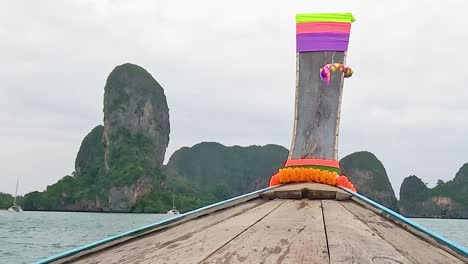 a wooden boat glides towards towering limestone formations under a cloudy sky.