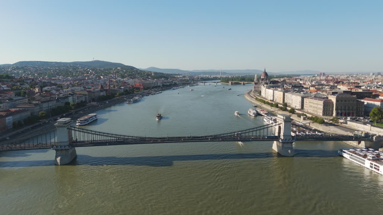 Aerial forward drone shot over Budapest’s Danube River, flying toward the Széchenyi Chain Bridge while ferry boats navigate the water below