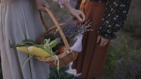 dos mujeres irreconocibles de pie entre el campo de lavanda con una cesta de alimentos y flores