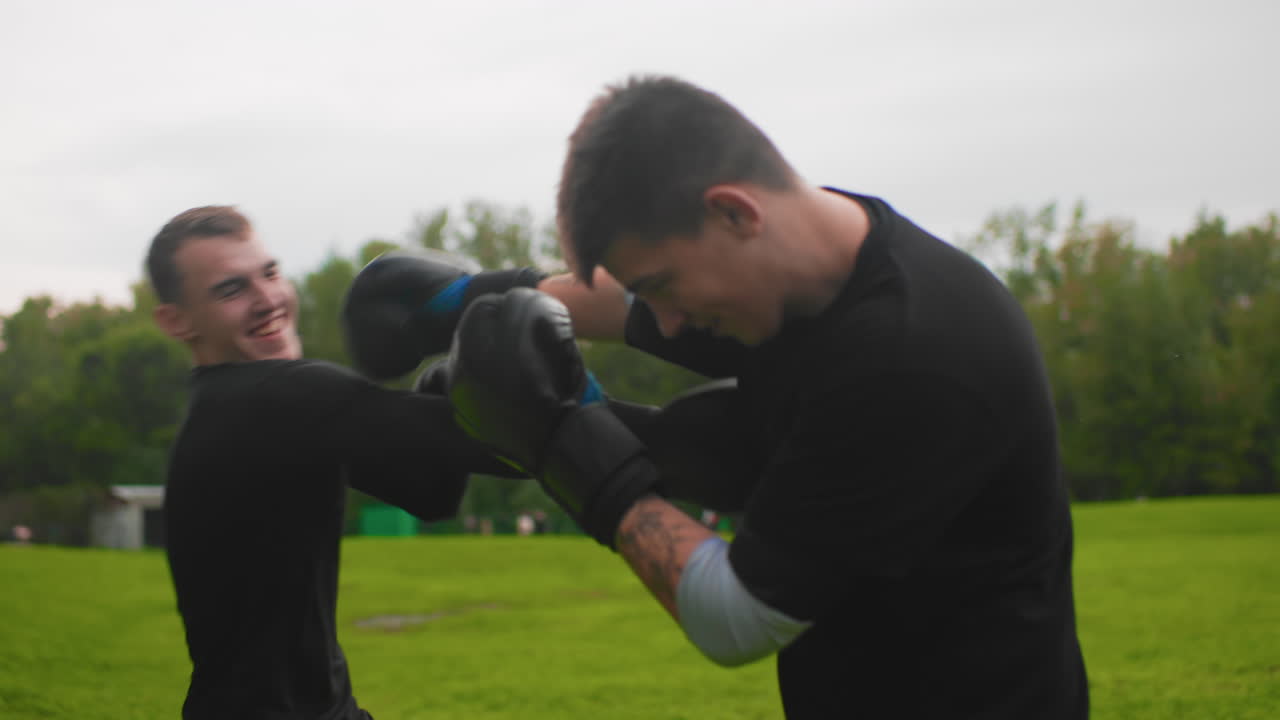 Homens brancos praticando sparring de forma divertida em um parque, trocas leves de luvas e jabs sorrindo entre amigos, sessão casual de fitness com céu nublado, ritmo fácil, brincadeiras, dicas de treinamento e energia calorosa capturada