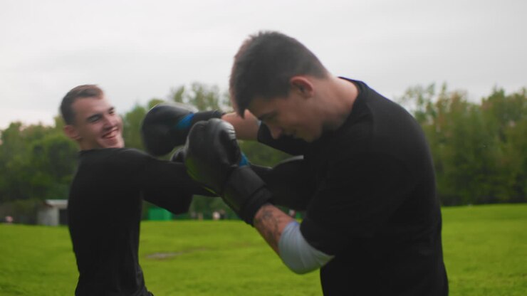white men playful sparring in park, lighthearted mitt exchanges and smiling jabs between friends casual fitness session with overcast sky, easy pace, banter, coaching tips, and warm energy captured