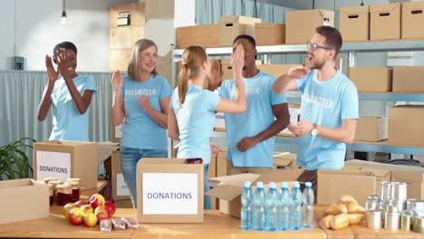 multiethnic group of volunteers standing in charity warehouse clapping hands and giving high five in charity warehouse