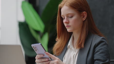 Una-Joven-Trabajadora-De-Oficina-Está-Usando-Un-Teléfono-Inteligente-Moderno-En-El-Lugar-De-Trabajo.-La-Mujer-Está-Revisando-Avisos-Y-Leyendo-Noticias.