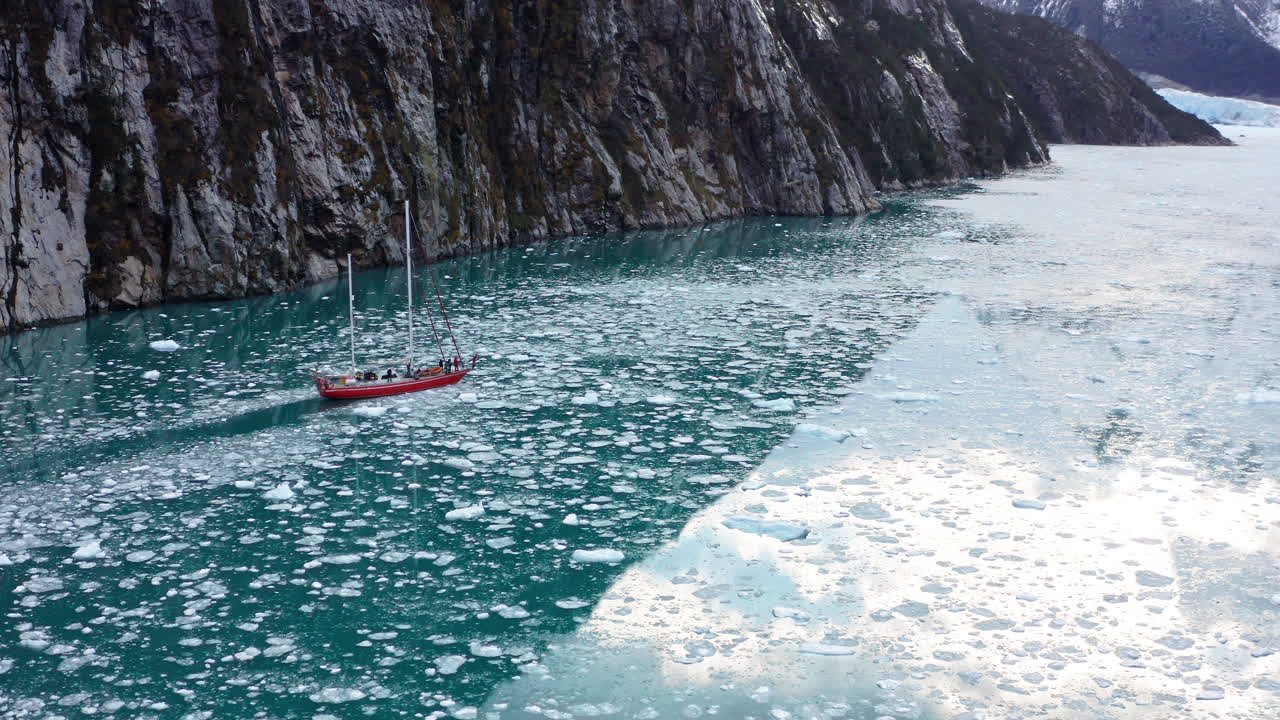 Sailboat slowly navigating between rugged coastal cliffs in Beagle Channel aerial crossing treacherous icy waters, aerial pullback