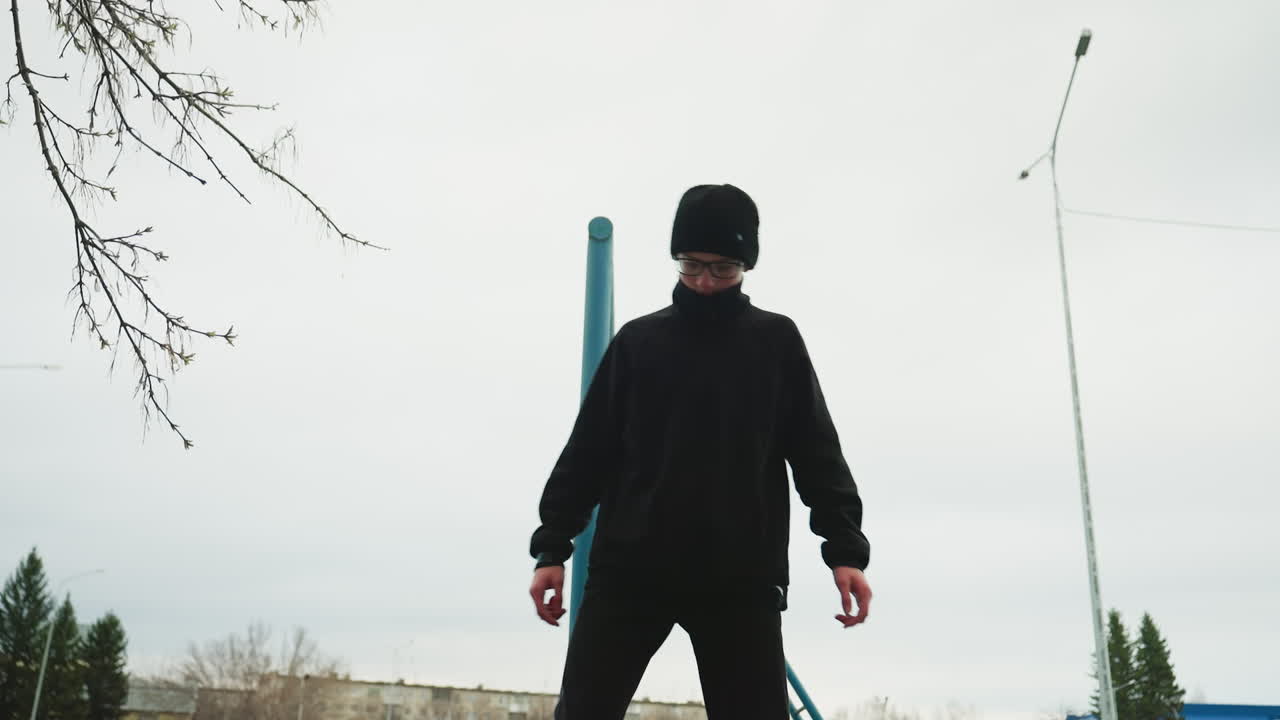A young boy dressed in a black outfit and glasses carefully walks on an elevated structure, while another boy in a gray sweater hangs on an iron pole in the background