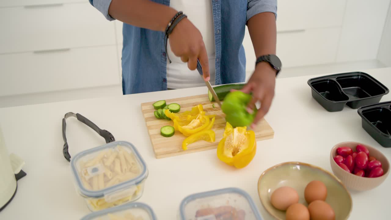Adult African American man placing pepper, cucumber on board slicing into prep trays in denim shirt