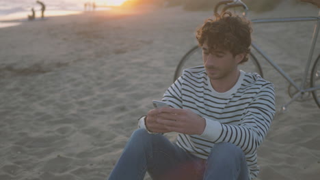 man sitting on the sand at the beach using the phone next to his bike