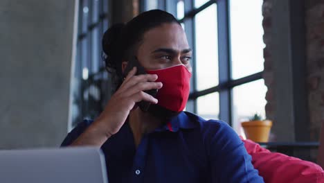 hombre afroamericano con máscara facial sentado en una cafetería hablando en un teléfono inteligente usando una computadora portátil