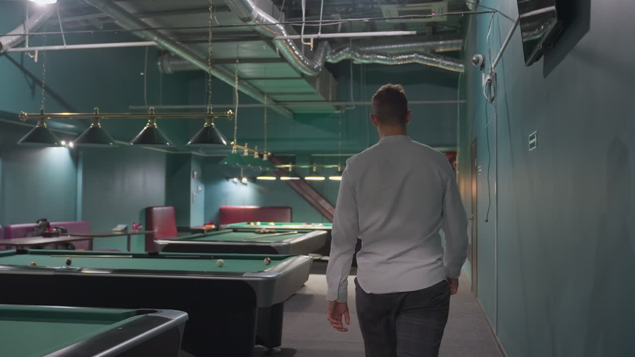 Back shot of young man in white shirt and grey trousers walking into billiard room with green pool tables. Overhead lamps illuminate stylish sports club setting with modern interior ambiance