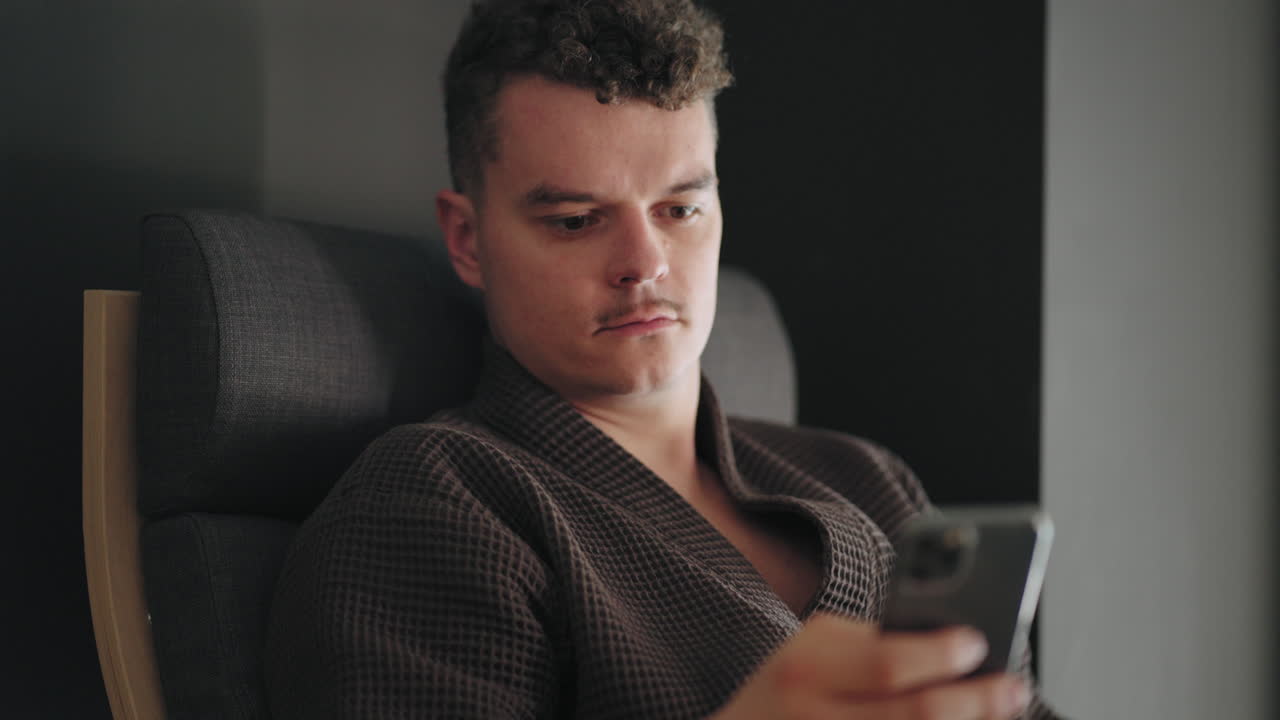 옷을 입은 성인 남자가 스파 센터에서 휴식을 취하고 치료 후 휴식을 취하고 스마트폰에서 뉴스와 이메일을 확인하고 있습니다.