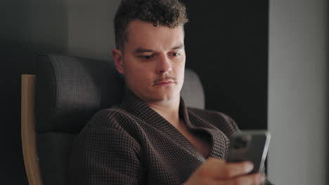 adult man in gown is relaxing in spa center resting after treatments checking news and email in smartphone